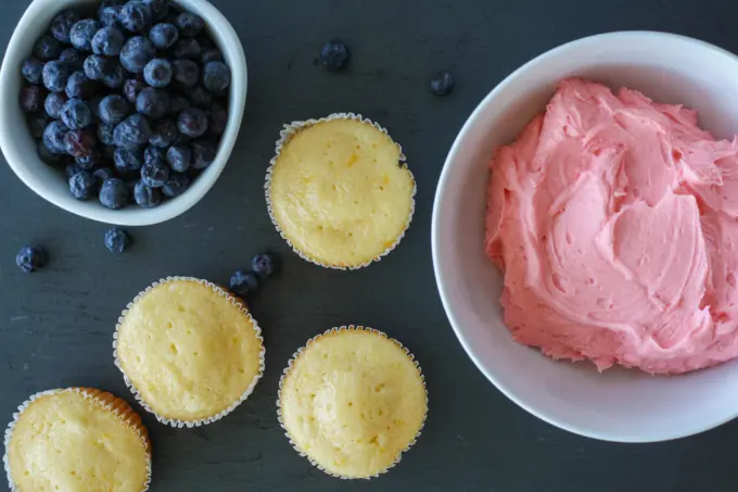 Blueberries in a bowl with unfrosted cupcakes and a bowl of pink buttercream.