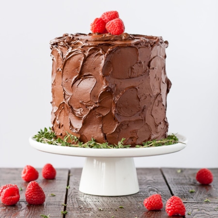 Chocolate covered cake on a white cake stand with fresh raspberries around it.