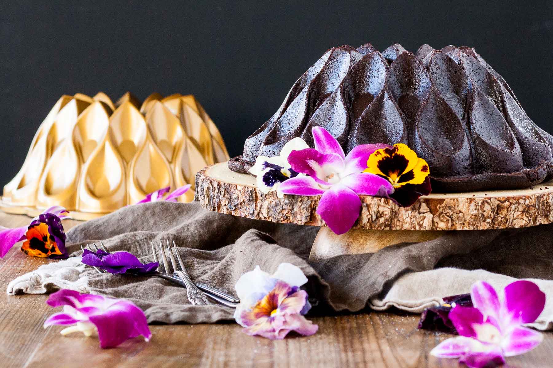 Chocolate Bundt cake on a wooden cake stand garnished with fresh edible flowers. Gold bundt pan in the background.