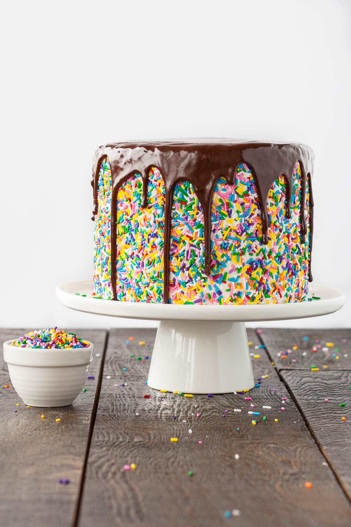 Funfetti cake with a chocolate ganache drip on a white cake stand. A bowl of sprinkles to the side.