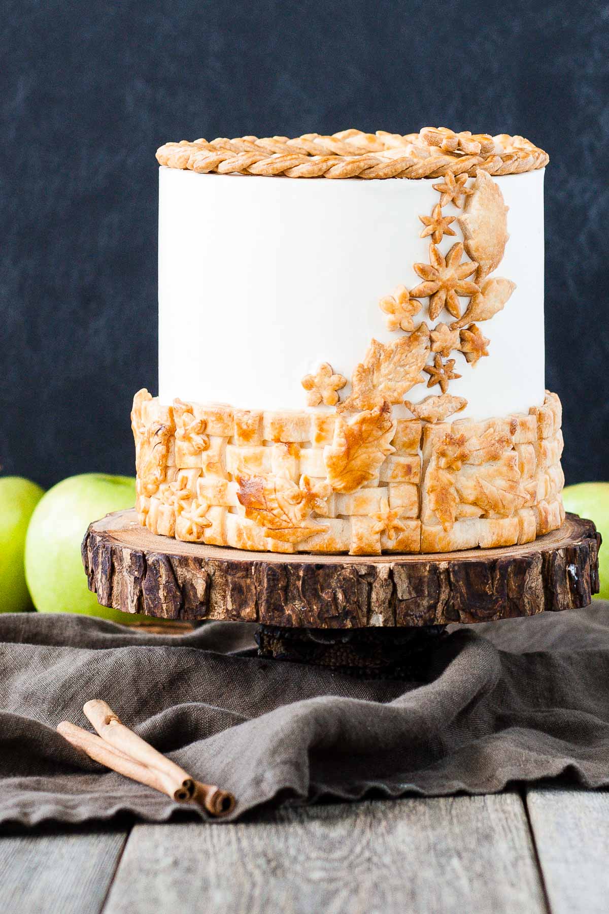 A cake sitting on top of a rustic wooden cake stand with apples in the background.