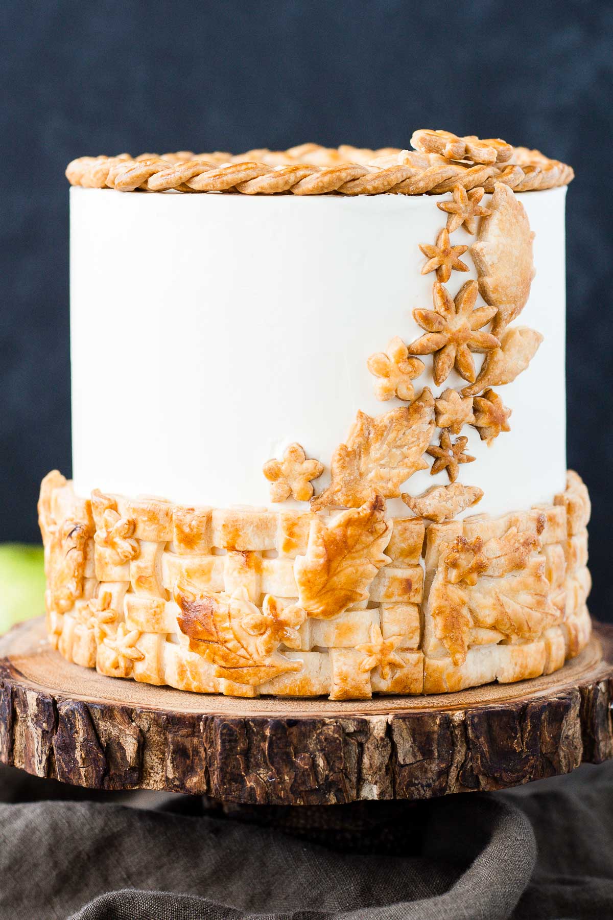 A cake sitting on top of a rustic wooden cake stand with apples in the background.