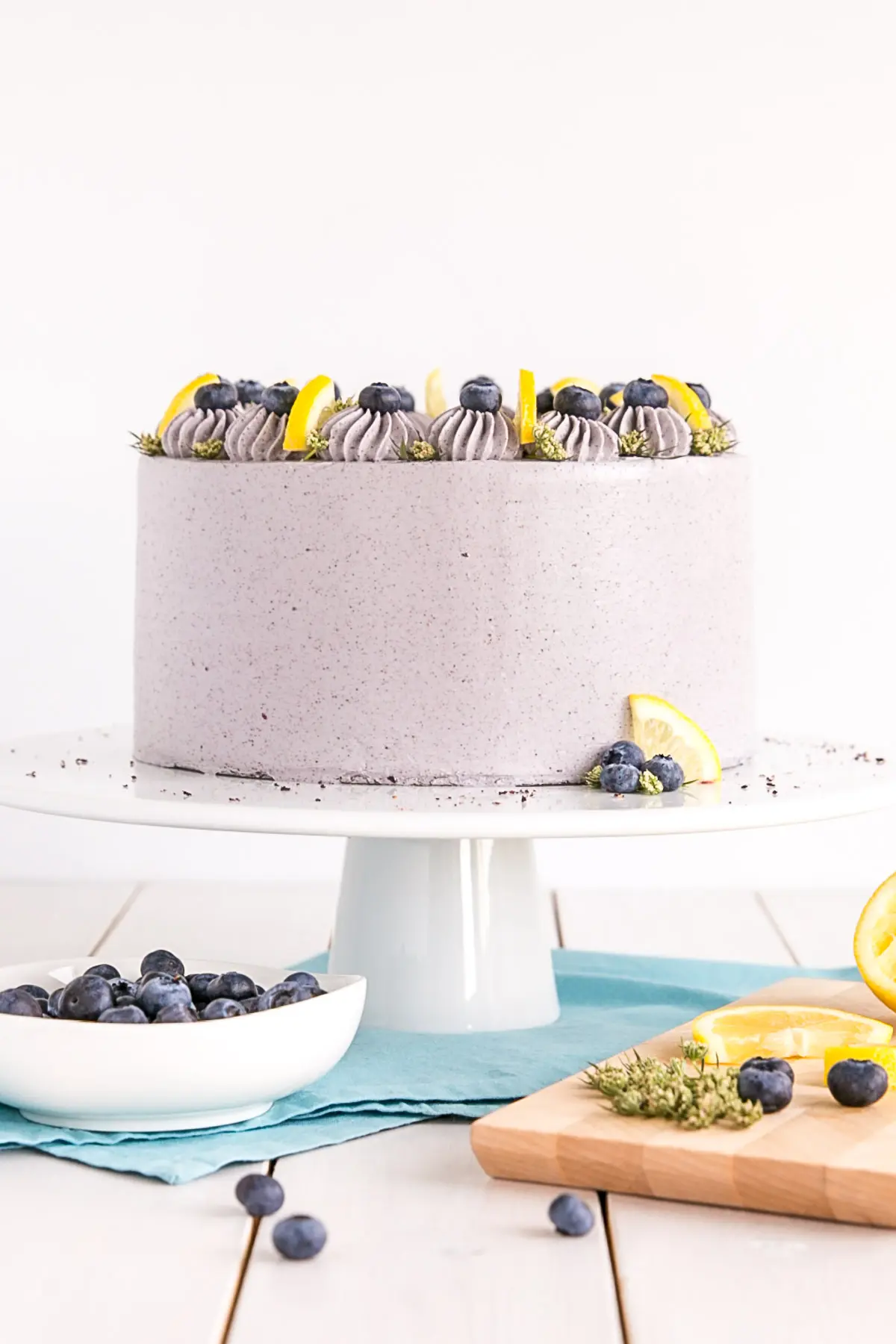 Cake on a white cake stand with a blue cloth underneath and a bowl of blueberries on the side.