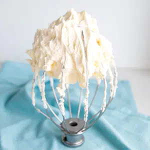 Buttercream on a whisk with a blue tea towel behind.