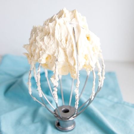 Buttercream on a whisk with a blue tea towel behind.