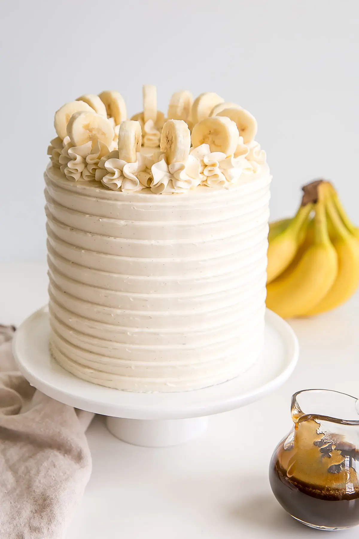 Bananas Foster cake with bananas in the background and fosters sauce in a small pitcher.