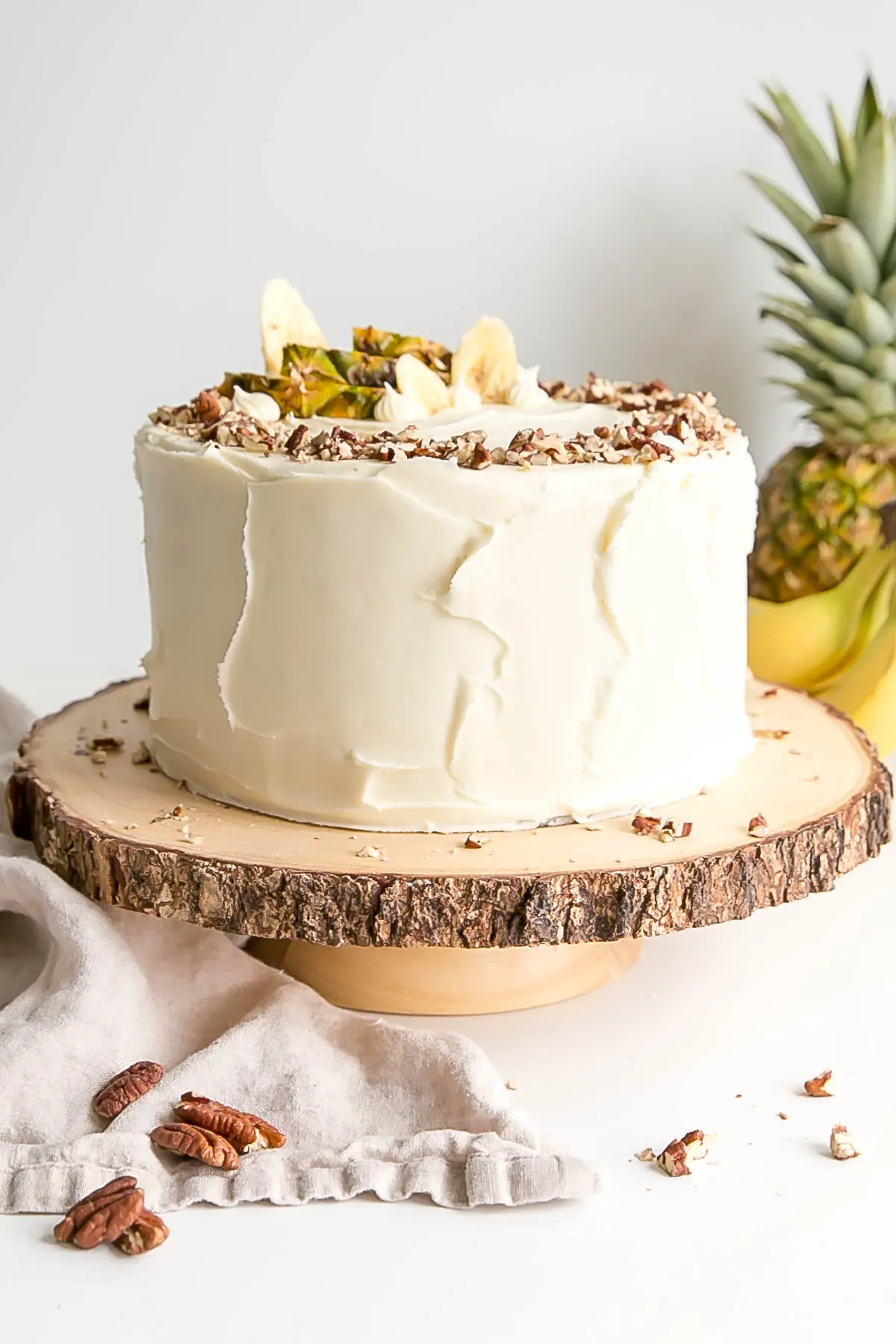 Hummingbird Cake on a rustic wood cake stand with a pineapple and bananas in the background.