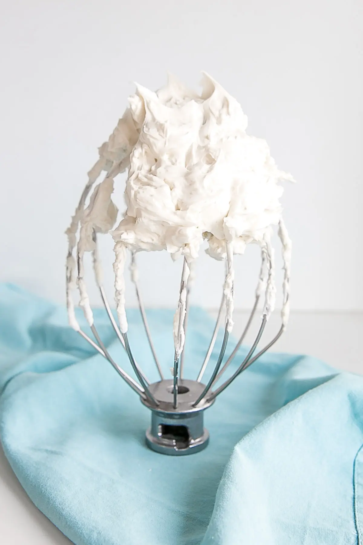 White buttercream on a stand mixer whisk on a blue cloth.